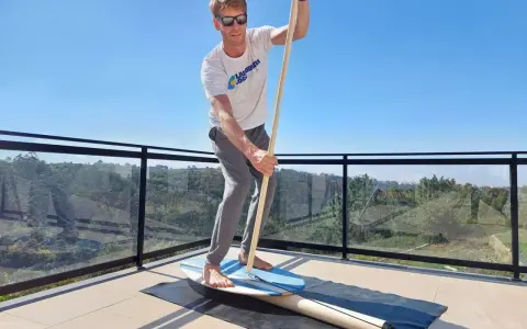 Mann übt Balance-Board-Training zuhause auf einer Terrasse. Er hält einen Paddelstock und balanciert auf einem Brett.