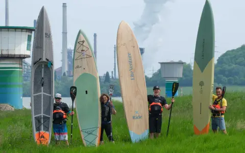 Fünf Personen mit SUP-Boards und Paddeln vor einer Industrieanlage. Die Boards zeigen fortgeschrittene SUP-Technik.