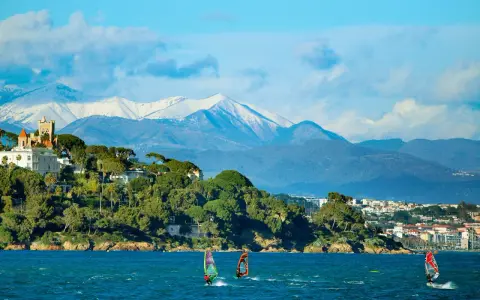 Windsurfen an der französischen Küste mit schneebedeckten Bergen im Hintergrund. Ein Schloss thront auf einem Hügel.