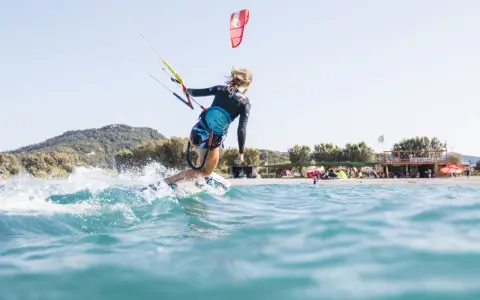 Eine Frau surft mit einem Kite auf Kos. Sie fährt auf einer Welle, Gischt spritzt. Im Hintergrund ist der Strand mit Bäumen und einem Gebäude zu sehen.