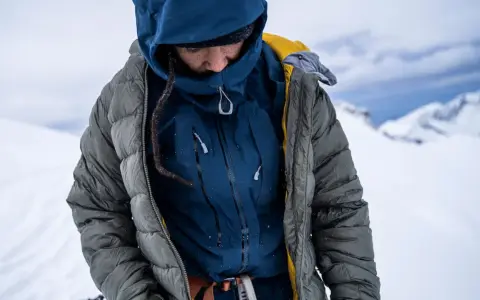Person in Daunenjacke im Schnee. Die Jacke ist grau und blau, mit gelben Akzenten. Perfekt für das Daunenjacke waschen nach einer Bergtour.