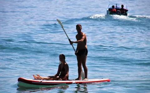 Ein Mann steht auf einem Stand Up Paddle Board auf dem Meer, während eine Frau sitzt. Im Hintergrund fährt ein Boot.