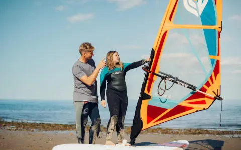 Vater erklärt Tochter das Windsurfen NRW. Sie stehen am Strand mit einem Segel und Board.
