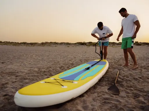 Zwei Männer bereiten ein SUP-Board am Strand vor. Einer pumpt es auf, fragt sich: "Sup, wieviel Bar?