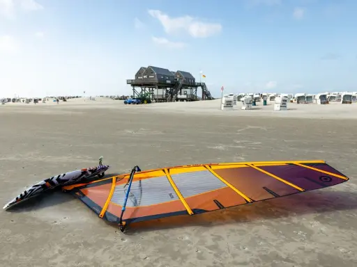 Windsurfen St. Peter-Ording: Segel und Brett liegen am Strand, bereit für den nächsten Ritt auf den Wellen. Strandkörbe und das markante Stelzenhaus im Hintergrund.