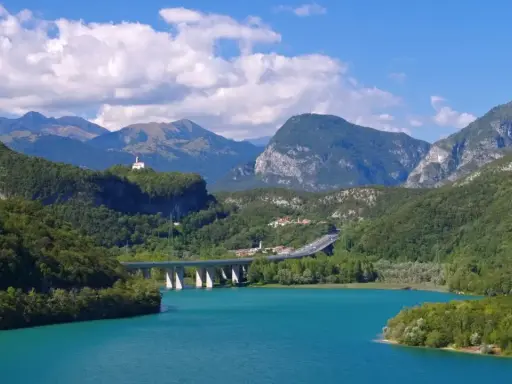 Türkises Wasser des Lago di Cavazzo umgeben von grünen Bergen. Eine Brücke überspannt den See, im Hintergrund eine kleine Kirche auf einem Hügel.