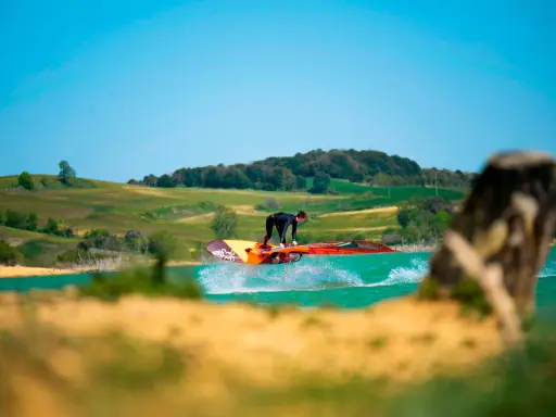 Windsurfer springt über das Wasser am Lac de Ganguise. Grüne Hügel und blauer Himmel im Hintergrund.