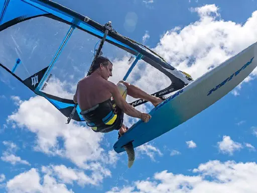 Ein Windsurfer mit einem Freeride Windsurfboard springt hoch in den blauen Himmel mit Wolken.