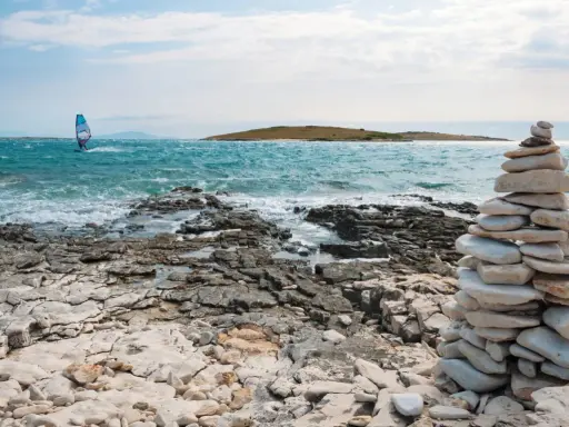 Ein Windsurfer gleitet über das türkisfarbene Meer vor Kroatien. Steinformationen säumen die felsige Küste.