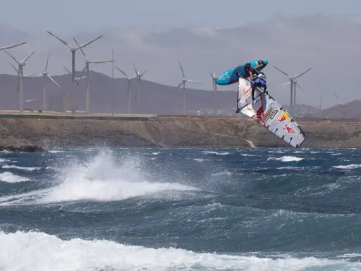 Windsurfer Britt Dunkerbeck springt hoch über den Wellen, im Hintergrund Windräder.