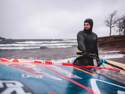 Mann in Neoprenanzug mit Windsurfbrett am Strand. Kaltes Wetter für Windsurfen in Deutschland.