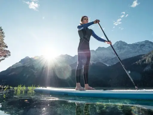 Eine Frau steht auf einem SUP-Board in Bayern, umgeben von Bergen und klarem Wasser.