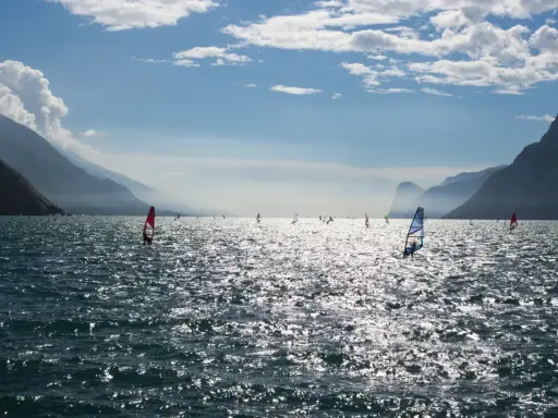 Windsurfer tummeln sich auf dem glitzernden Gardasee vor einer Bergkulisse.