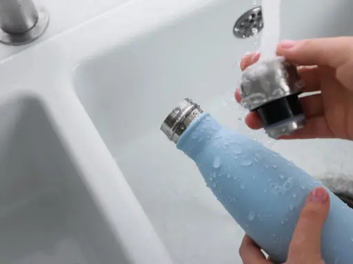 Hände reinigen eine hellblaue Trinkflasche unter fließendem Wasser im Waschbecken.
