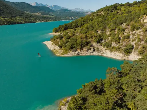 Türkisfarbener Lac de Monteynard mit einem Windsurfer, umgeben von bewaldeten Hügeln und Bergen.