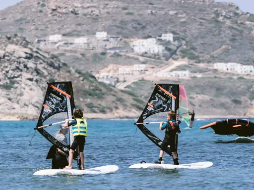 Kinder lernen naxos surfen. Zwei junge Windsurfer üben auf dem Meer, im Hintergrund weiße Häuser am Hang.