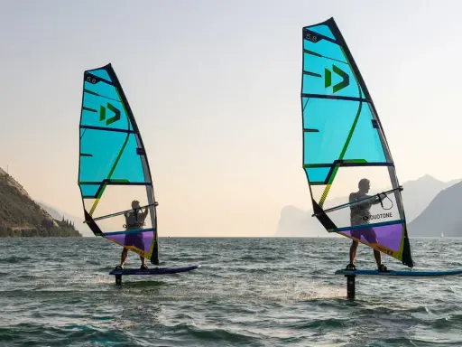 Zwei Personen beim Windsurfen in Italien. Die türkisfarbenen Segel sind im Gegenlicht zu sehen, im Hintergrund Berge.