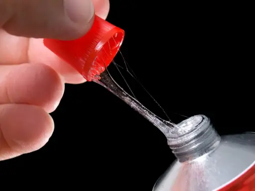 Ein Finger hält die rote Kappe eines Tubenklebers, aus dem zäher, glitzernder Kleber in Fäden heraustropft.