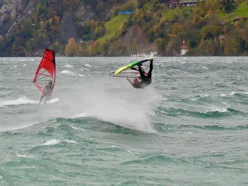 Windsurfer vollführt Sprung am Starnberger See. Ein weiterer Surfer mit rotem Segel im Hintergrund.