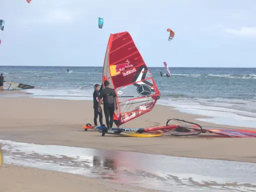 Windsurfer bereiten sich am Strand von Pozo Izquierdo, Gran Canaria, auf das Wasser vor. Mehrere bunte Segel und Boards liegen am Sand.