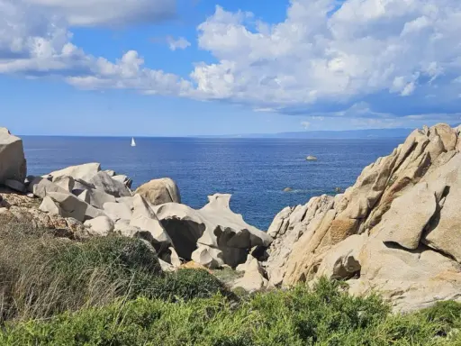 Felsformationen am Capo Testa mit Blick auf das blaue Meer und einen einsamen Segler unter bewölktem Himmel.