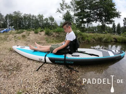 Junge sitzt auf SUP-Board, um den SUP Sitz zu befestigen. Das Board liegt am Ufer.