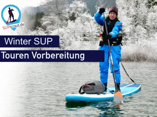 Person im blauen Neoprenanzug bereitet sich auf eine Winter SUP Tour vor. Die Landschaft ist mit Schnee bedeckt.