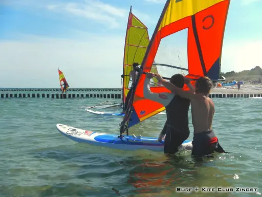 Zwei junge Männer bereiten sich auf das Windsurfen an der Ostsee vor. Ein weiterer Surfer ist in der Ferne zu sehen.