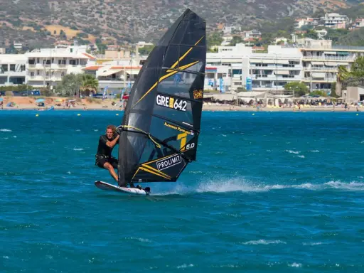 Windsurfer mit schwarzem Segel und gelben Akzenten auf türkisfarbenem Wasser vor einer Küste mit Gebäuden und Bergen. Surfen in Griechenland.