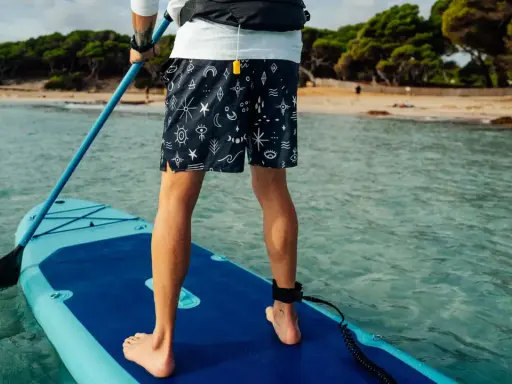 Person übt SUP Training auf einem Board im türkisfarbenen Wasser. Im Hintergrund eine sandige Bucht mit Bäumen.