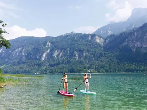 Zwei Frauen genießen Stand-Up-Paddling auf einem ruhigen See vor einer Bergkulisse. Perfekte sup spots für einen entspannten Tag.