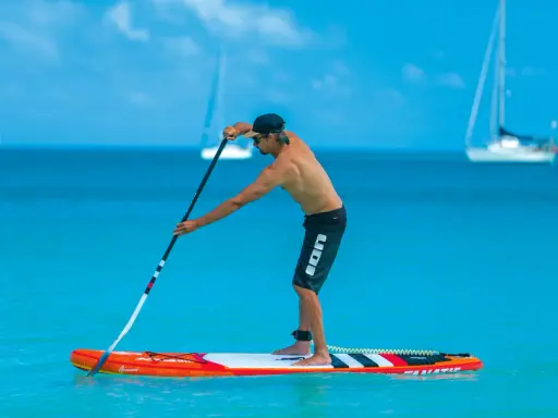 Mann stellt sein SUP Paddel ein, während er auf einem Board im türkisfarbenen Wasser steht. Segelboote sind im Hintergrund zu sehen.