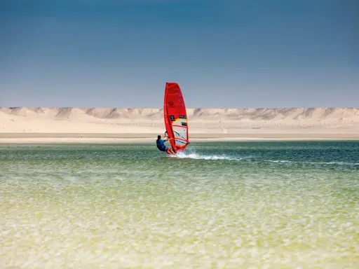 Windsurfer mit rotem Segel gleitet über das Wasser bei Dakhla, Marokko, vor einer Wüstenlandschaft.