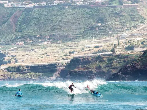 Surfer auf La Gomera reiten eine Welle, während im Hintergrund terrassierte Hänge und Häuser zu sehen sind.
