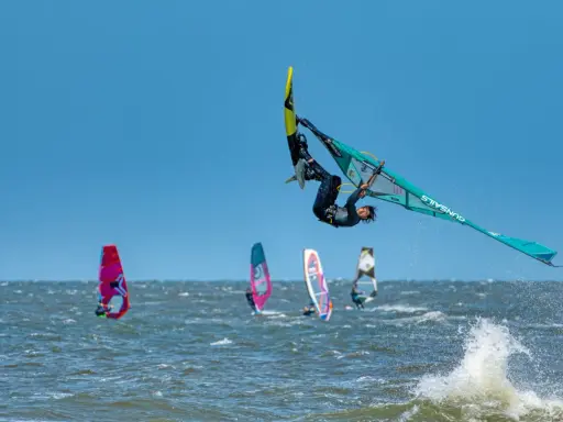 Windsurfer vollführt spektakulären Sprung über den Wellen. Mehrere andere Windsurfer sind auf dem Wasser zu sehen, was auf beliebte Windsurfing Spots hindeutet.