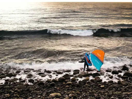 Ein Wingfoiler steht am steinigen Strand, bereit für den **shore break**. Die Sonne wirft ein warmes Licht auf die Wellen.