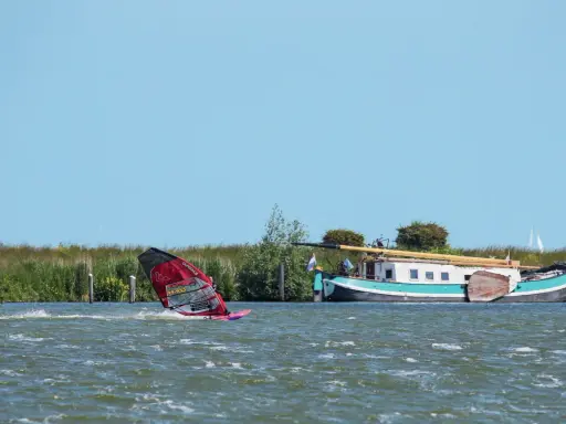 Windsurfen Holland: Ein Surfer gleitet über das Wasser, während ein Hausboot am Ufer liegt.
