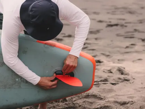Mann mit Hut befestigt die rote Finne an einem aufblasbaren SUP-Board. Gute SUP-Pflege beginnt mit dem richtigen Zubehör.