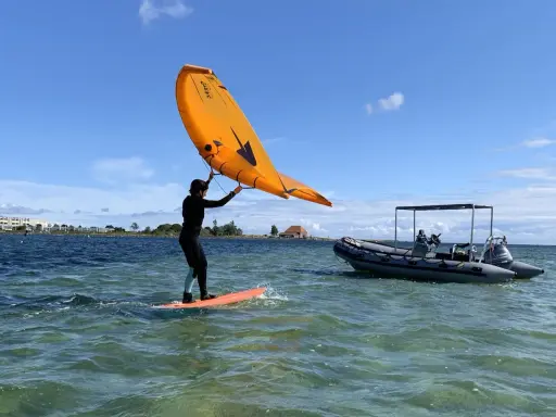 Person auf einem Board mit einem orangen Flügel, der über dem Wasser schwebt. Ein Schlauchboot liegt daneben. Foil surfen ohne Segel.