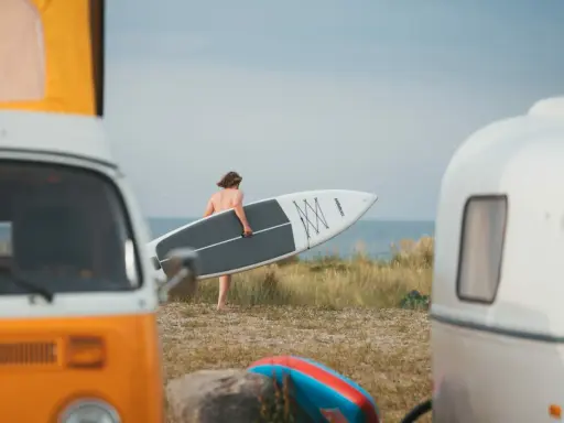 Mann mit SUP-Board am Strand von Fehmarn, bereit für die Wellen. Wohnmobile im Vordergrund.