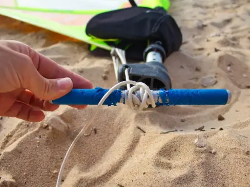 Hand hält blauen Griff, um das Segel zu spannen. Windsurf-Ausrüstung liegt im Sand.