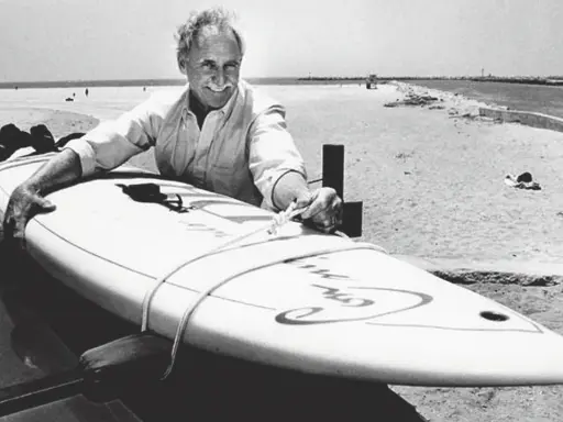 Jim Drake, ein Pionier des Surfens, lächelt stolz neben seinem Surfbrett am Strand.