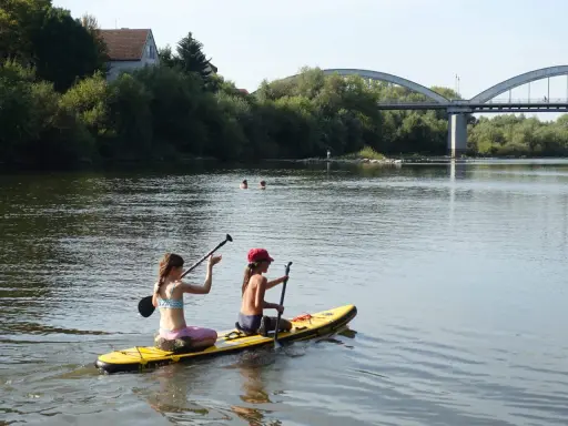 Zwei Mädchen genießen das Stand Up Paddling in Heidelberg. Im Hintergrund eine Brücke und Bäume.