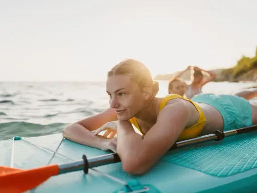 Junge Frau entspannt auf SUP-Board im Wasser. Ob Stand up paddling schwer ist, fragt sie sich lächelnd, während Freunde im Hintergrund planschen.