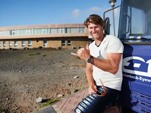 Bekannte deutsche Windsurfer: Ein Mann mit Sonnenbrille und Shaka-Geste lehnt an einem blauen Bus vor dem Centro Internacional de Windsurfing.