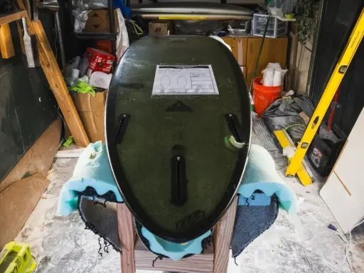 Surfboard liegt auf Ständer, bereit für die **Surfboard Reparatur**. Werkzeuge und Materialien im Hintergrund.