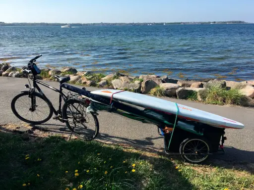 Fahrradanhänger mit Surfbrett und Paddel am Seeufer, bereit für ein Abenteuer auf dem Wasser.