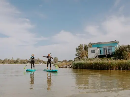 Zwei Frauen auf SUP-Boards am Makkum Strand, mit einem modernen Haus im Hintergrund.