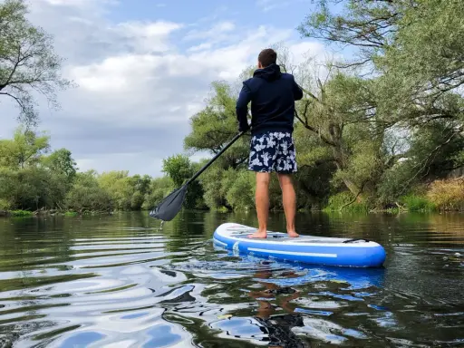 Mann auf SUP-Board auf ruhigem Fluss, umgeben von grünen Bäumen. Perfekt, um zu sehen, wo man stand up paddeln kann.