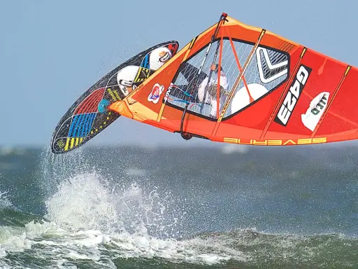 Lina Erpenstein fliegt mit ihrem Windsurf-Equipment über die Wellen. Das orangefarbene Segel und das bunte Board sind in der Luft.